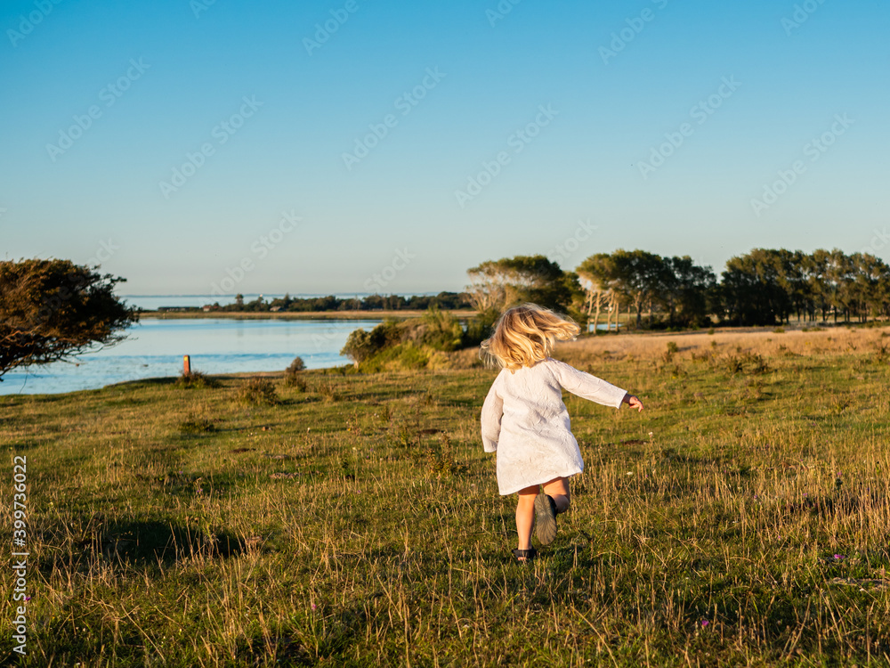 Blond girl 4 years old in white summer dress running and dancing on a field in sunlight and blue sky above