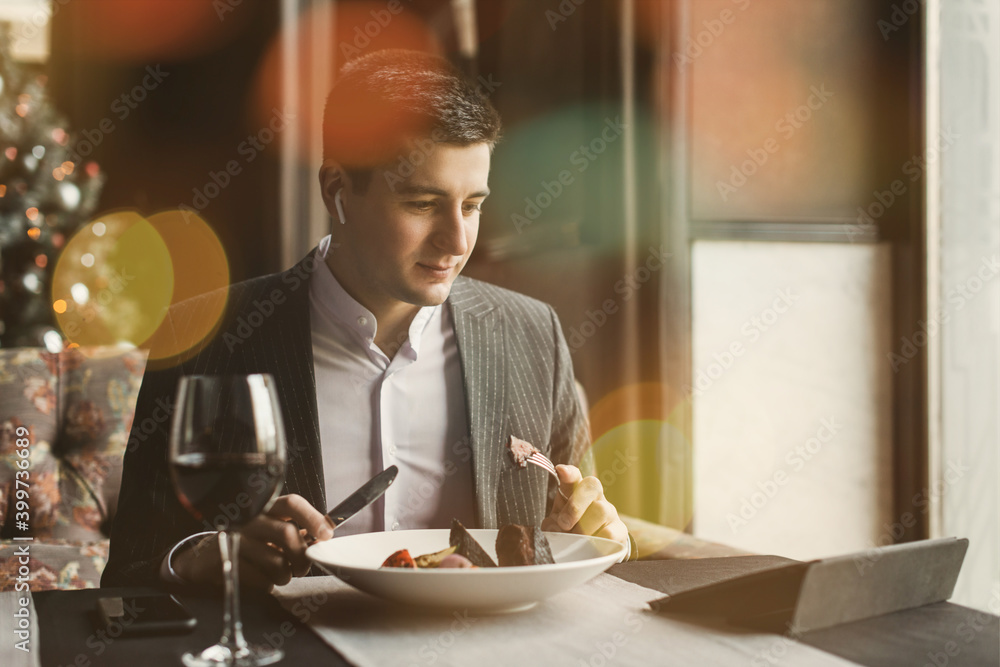 A young and attractive man uses his phone while eating veal tenderloin in ashes with grilled vegetables in an indoor restaurant A young and attractive man uses his phone while eating veal tenderloin in ashes with grilled vegetables in an indoor restaurant