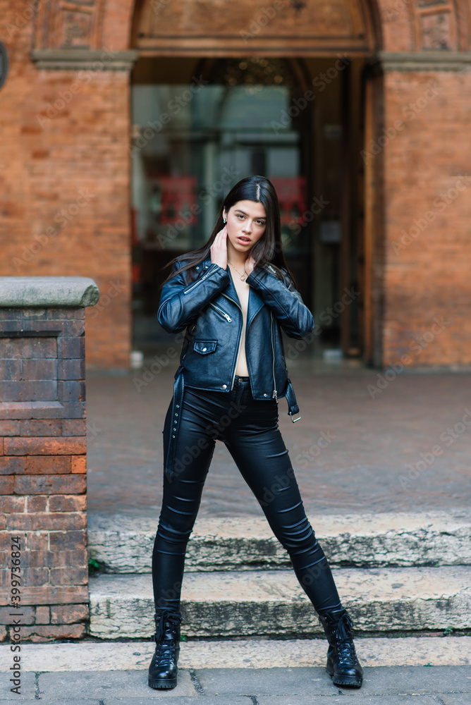 Portrait of beautiful young brunette woman in stylish black leather jacket, smiling on urban back