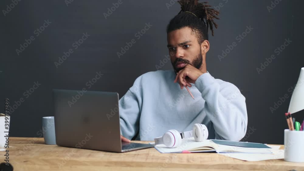 A serious young african american man is working with his laptop computer sitting at the table over a grey wall at home A serious young african american man is working with his laptop computer sitting at the table over a grey wall at home