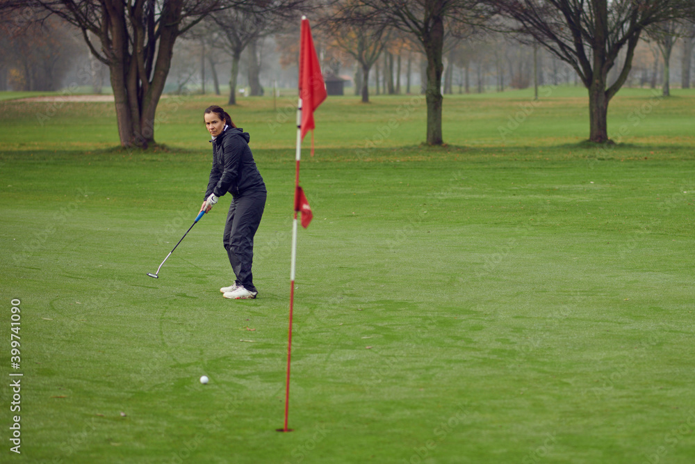 Obraz premium Middle-aged fit woman golfer putting for the hole on the green viewed from the flag as her golf ball approaches on a cold windy misty day