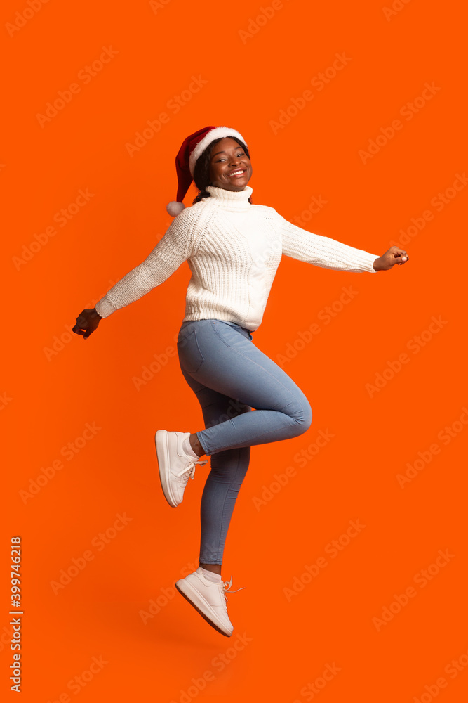Female Santa. Joyful Black Woman In Xmas Hat Jumping Over Orange Background