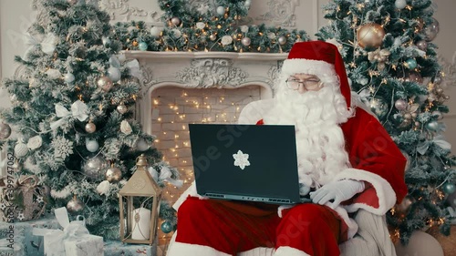 Santa Claus is typing on a laptop. Santa is sitting in a chair with a laptop and typing a response to an email.