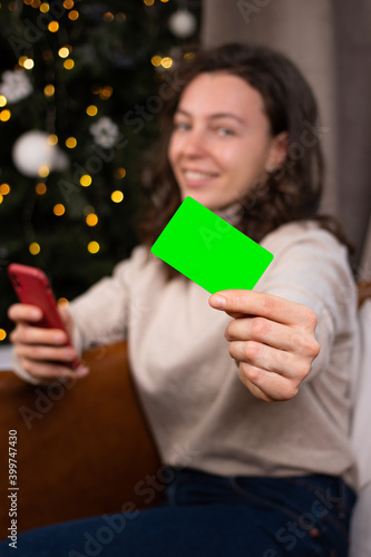 Blurry smiling woman holding in front of her blank credit bank card for mock up, looking at camera, using smartphone and sitting near Christmas tree at home. New Year online shopping. Vertical photo