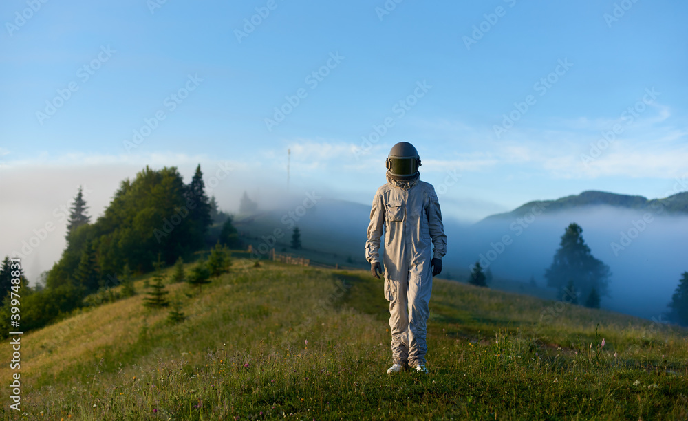 Portrait of spaceman wearing white space suit and helmet walking alone sunny green mountain glade in the morning, foggy hills and blue sky on background. Concept of astronautics and nature