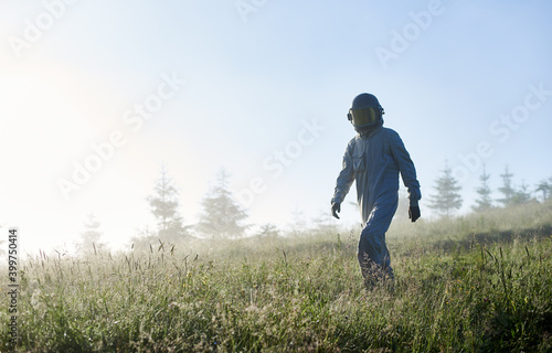 Cosmonaut in space suit walking down the meadow with lush grass. Male astronaut in helmet strolling down grass field with light blue sky on background. Concept of astronautics, exploration and nature. © anatoliy_gleb
