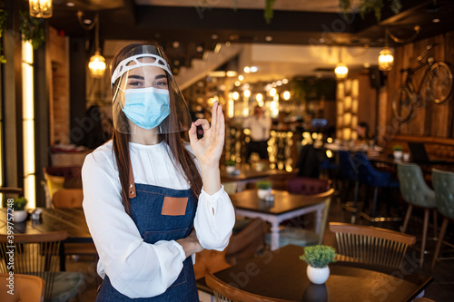 Portrait attractive waitress wear face mask and face shield showing OK sign with indoor restaurant background. New normal restaurant concept. Beautiful waitress working at a cafe