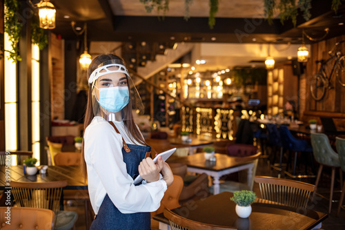 Female waitress wearing protective face mask and visor while holding touchpad and reopening sidewalk cafe during COVID-19 epidemic. New normal restaurant concept.