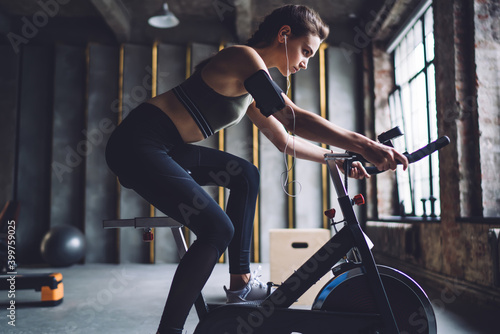 Serious caucasian female in activewear sitting on exercising bike ready for reaching good results in cardiom determined woman listening online radio via sound accessory slimming and losing weight