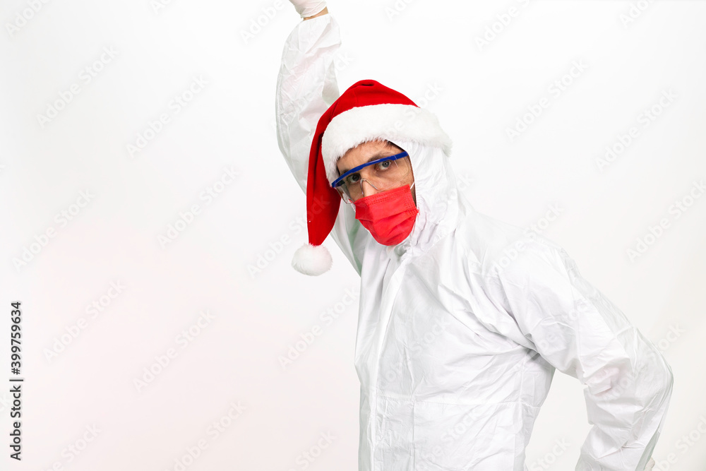 Health worker wearing protective clothing. He wears a santa hat on his head. He has a red corona mask on his face.