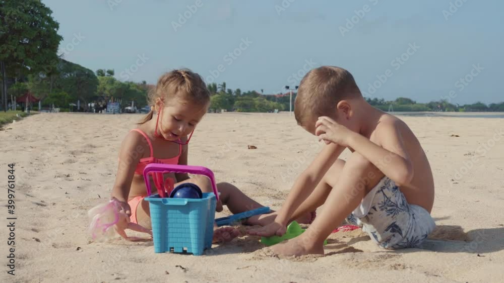 Lockdown of little Caucasian girl wearing pink swimsuit and her elder brother sitting together on sand on seacoast and playing with toys Lockdown of little Caucasian girl wearing pink swimsuit and her elder brother sitting together on sand on seacoast and playing with toys