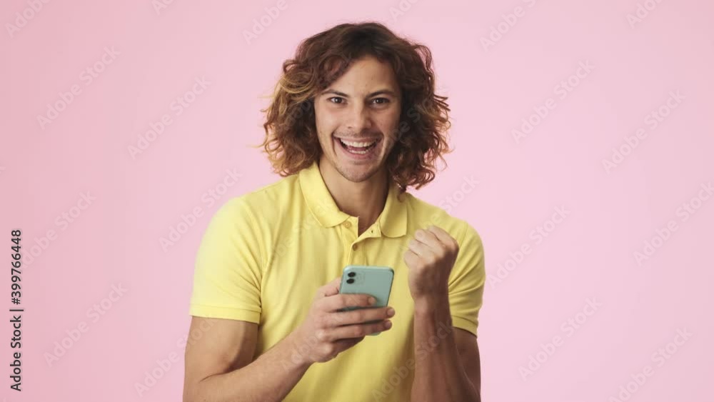 A smiling curly young man is doing winner gesture while using his smartphone standing isolated over a pink background in the studio A smiling curly young man is doing winner gesture while using his smartphone standing isolated over a pink background in the studio