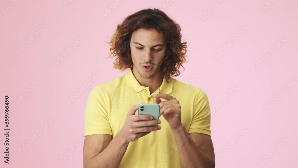 A smiling curly young man is doing thumb-up gesture while using his smartphone standing isolated over a pink background in the studio A smiling curly young man is doing thumb-up gesture while using his smartphone standing isolated over a pink background in the studio