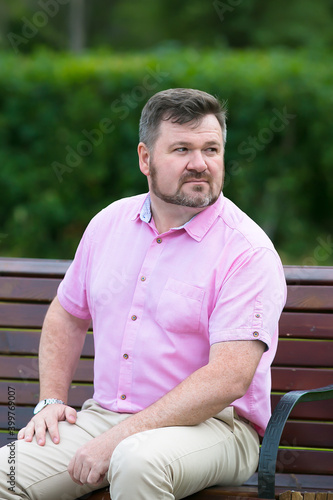 An adult man with a beard is bored in the park on a bench.