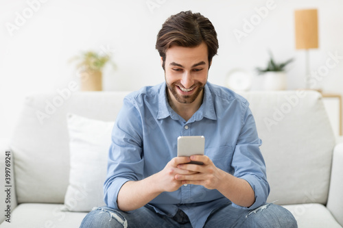 Excited Man Using Phone Texting Sitting On Sofa At Home