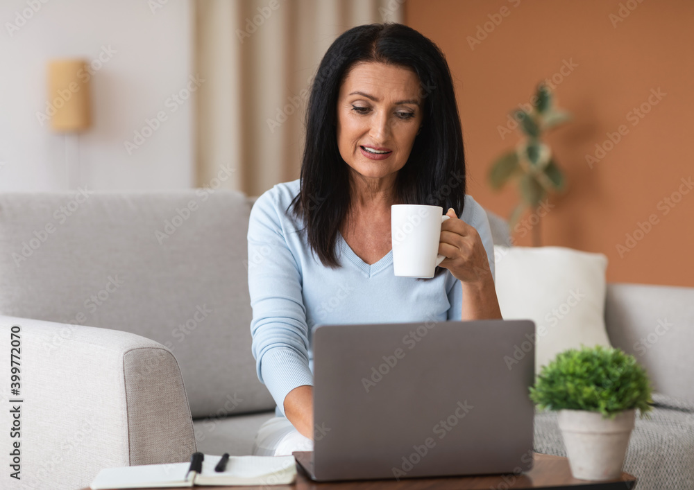 Mature woman sitting on sofa drinking coffe working on computer Mature woman sitting on sofa drinking coffe working on computer