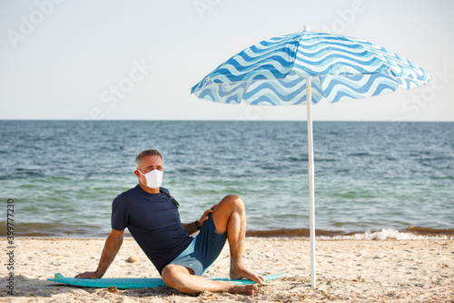 Uomo cinquantenne con mascherina protettiva sta in spiaggia sotto l'ombrello vicino al mare photo