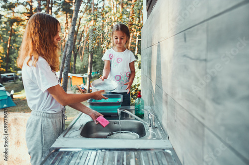 Teenager girl washing up the dishes pots and plates with help her younger sister in the outdoor kitchen during vacations on camping. Camp life. Sisters spending time and working together