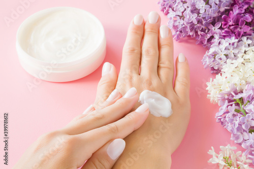 Young perfect adult woman hands using white moisturizing cream. Care about nails and clean  soft  smooth body skin. Beautiful branches of fresh  colorful lilac flowers. Closeup. Point of view shot.