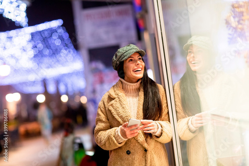 Beautiful young woman with mobile phone by the shop window at Christmas time