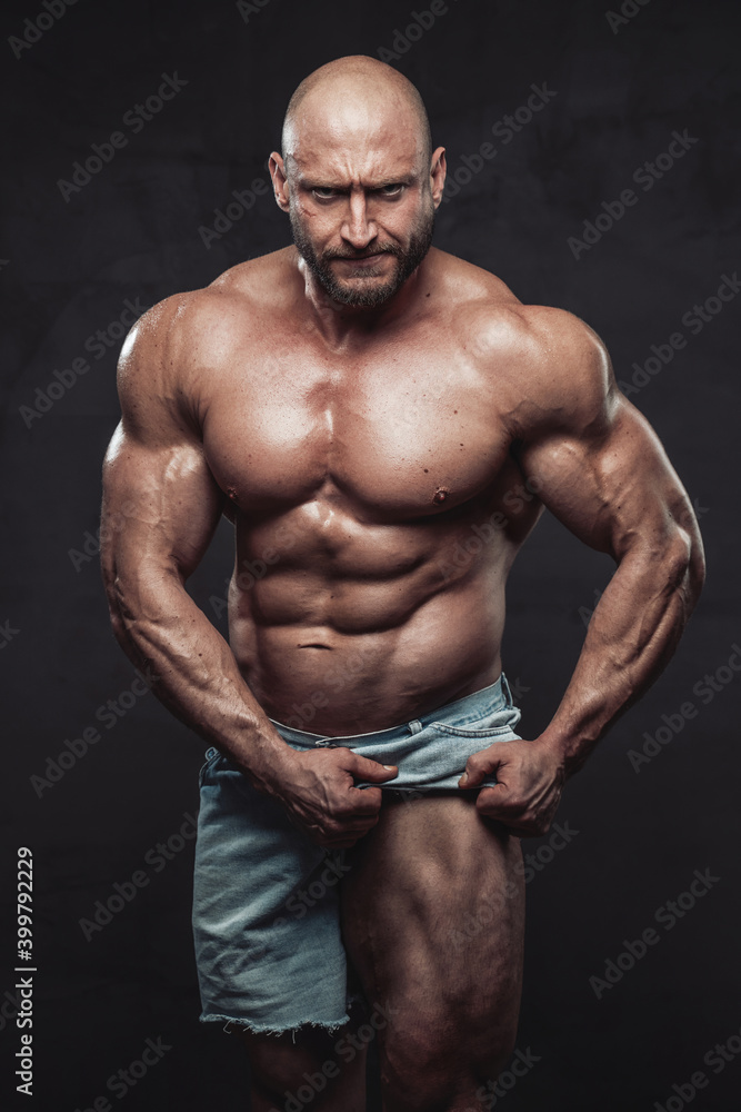 Strong and shirtless bodybuilder with beard and bald head poses in dark background pulling his shorts and showing his muscular leg looking at camera with angry face.