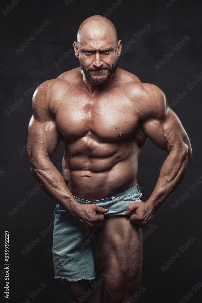Strong and shirtless bodybuilder with beard and bald head poses in dark background pulling his shorts and showing his muscular leg looking at camera with angry face. Strong and shirtless bodybuilder with beard and bald head poses in dark background pulling his shorts and showing his muscular leg looking at camera with angry face.
