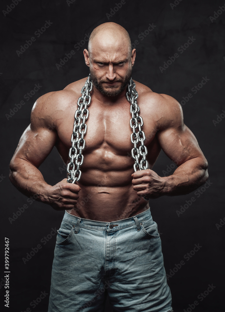 Strong and sportive man with muscular topless build poses holding heavy steel chains in dark background looking at camera with serious face.