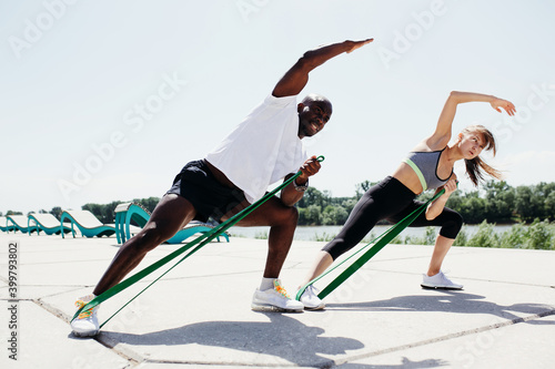 Afroamerican man in white t-shirt and black shorts with tape expander doint exercises with women in grey top and black pants on fresh air and sunny weather.