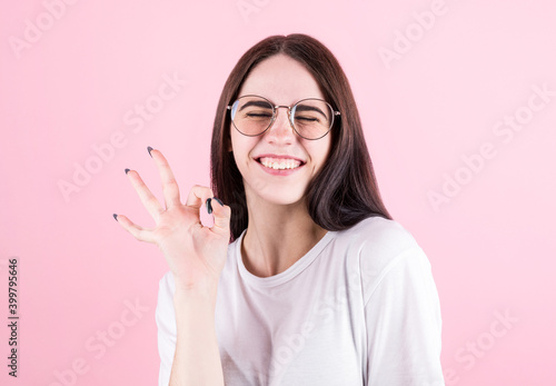 Close-up photo of funny young brunette woman in white t-shirt showing OK gesture, looking at camera, isolated on pink background