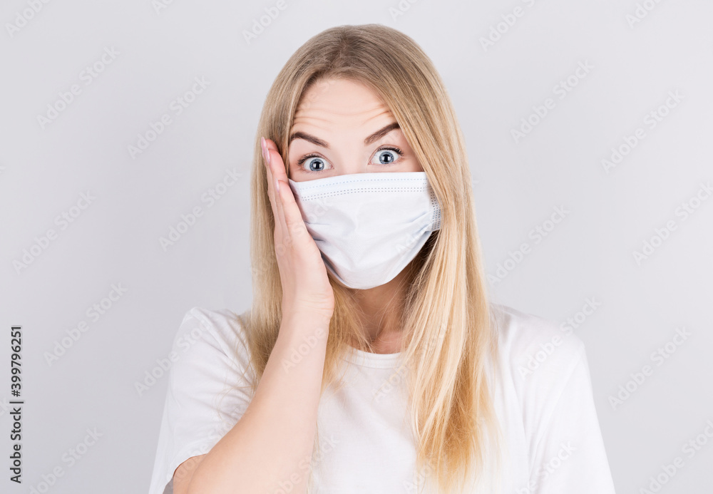 Beautiful caucasian young woman in white t-shirt with disposable face mask surprised and looking at the camera with his hand to his face. Protection versus viruses and infection. Beautiful caucasian young woman in white t-shirt with disposable face mask surprised and looking at the camera with his hand to his face. Protection versus viruses and infection.