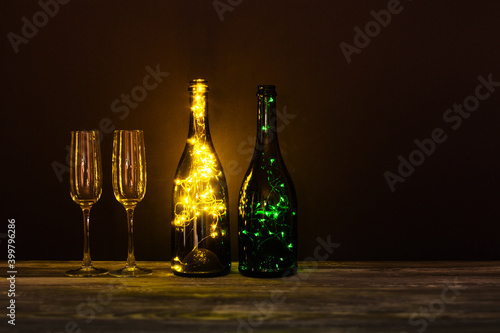 Festive romantic postcard. Shining champagne wineglasses, wooden table on dark background. Cozy still life composition bottles decorated garlands. Christmas Valentines day holiday greeting card.