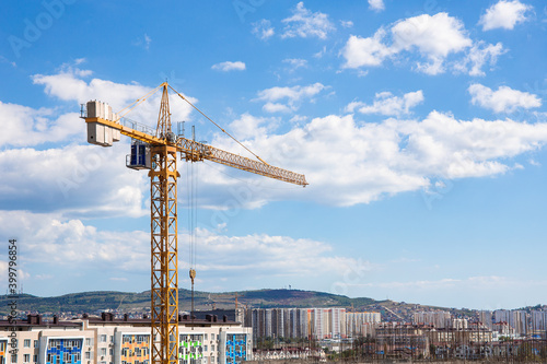 Yellow construction crane or tower with a cabin at height, against the backdrop of a clear blue sky and cityscape