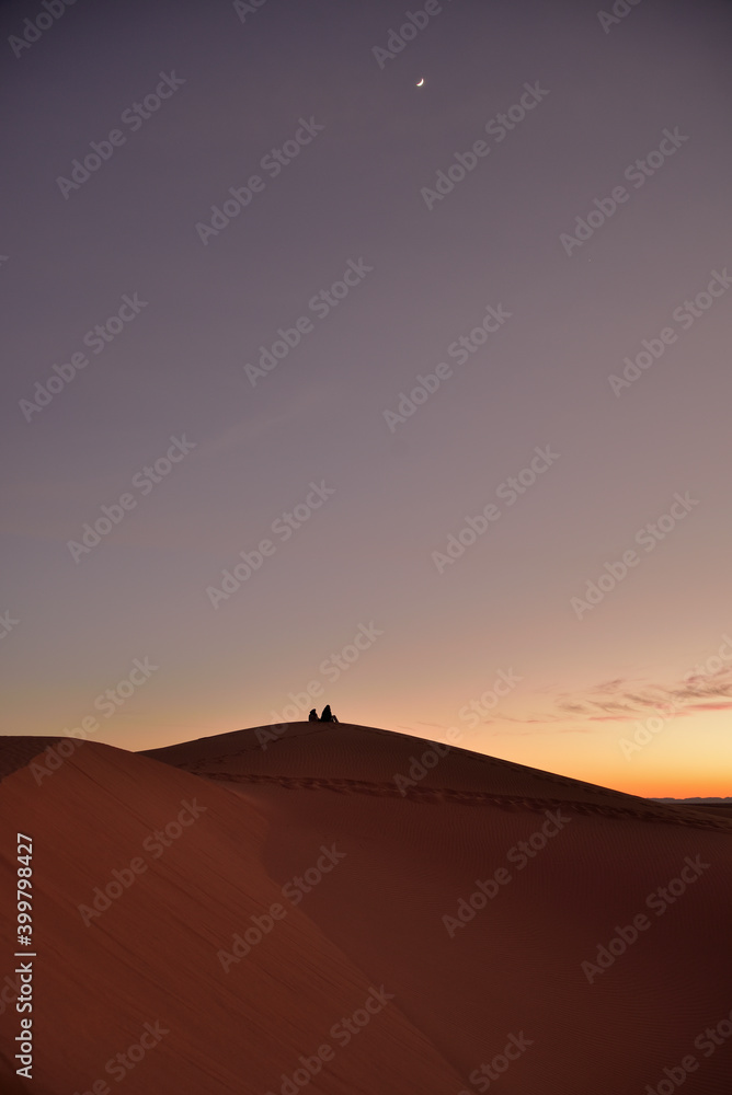 Die Silhouette von zwei Personen auf einem Sandberg in der Wüste, der den Sonnenuntergang und einen Halbmond am Himmel beobachtet