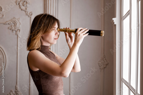A woman in a tight-fitting dress looks through a spyglass near the window from the room.