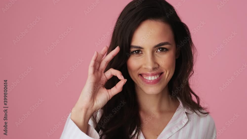 A smiling young woman wearing pajamas is doing ok gesture standing isolated over a pink background in the studio A smiling young woman wearing pajamas is doing ok gesture standing isolated over a pink background in the studio