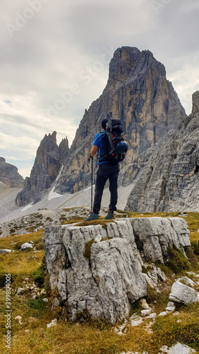 Man with big backpack and sticks, hiking in high Italian Dolomites. There are many sharp peaks in front of him. He is standing on a big boulder. Lots of lose stones and landslides. Sunny day. Outdoor © Chris