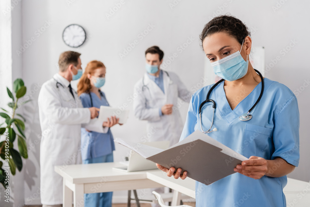 African american nurse in medical mask looking at paper folder with blurred colleagues working on background African american nurse in medical mask looking at paper folder with blurred colleagues working on background