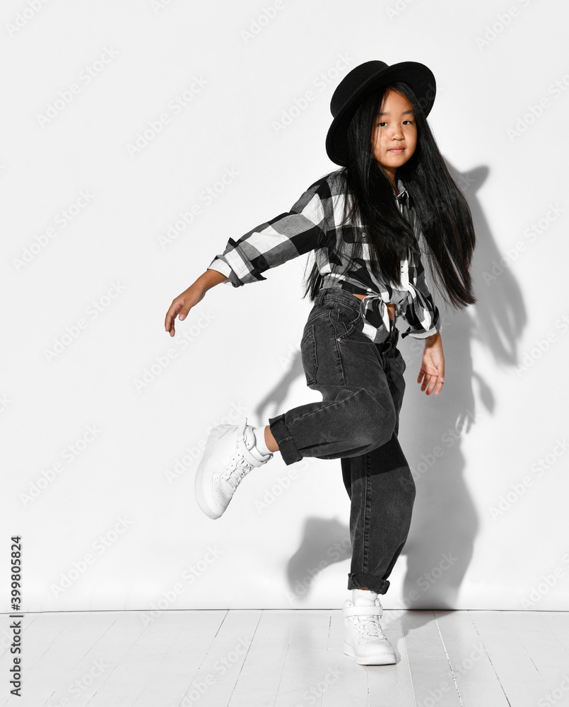Naklejka premium Portrait of an Asian girl with black hair who dances and has fun on a white background. The child wears a black hat, plaid shirt, jeans and looks into the camera. Children's fashion and style.