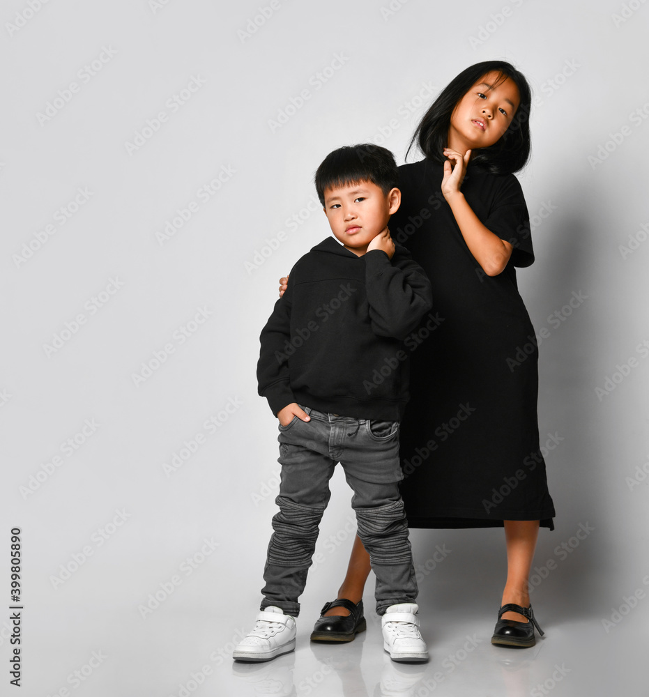 Obraz premium Portrait of two beautiful Asian children posing in front of the camera in the studio against a white background. A cool boy in a black hoodie and jeans with an older sister in a black dress is fun.