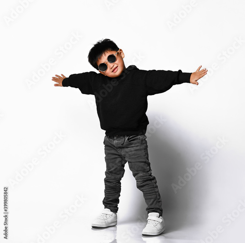 Portrait of a cute little boy in stylish clothes and sunglasses, arms outstretched and having fun at the white studio wall. Children's fashion concept.