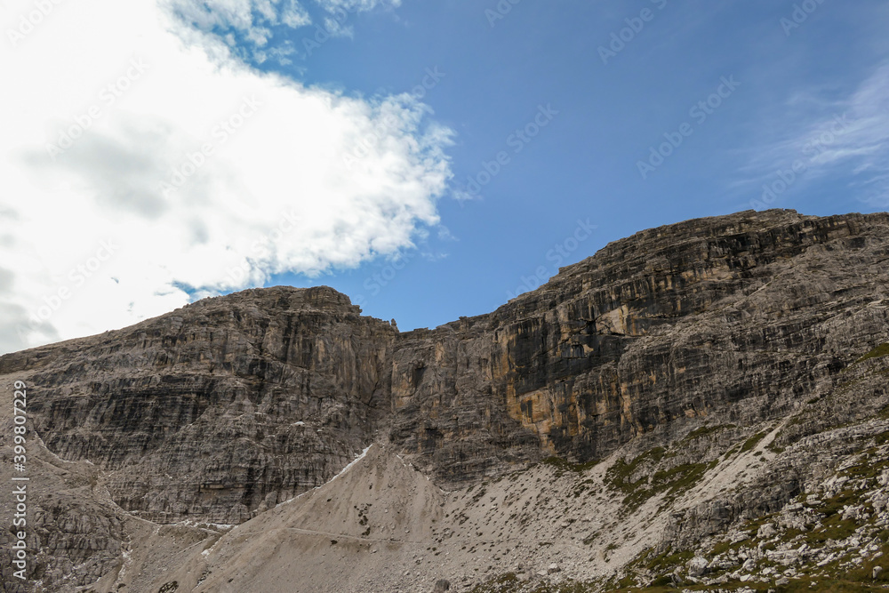 A close up view on a massive, high and desolated mountain wall in Italian Dolomites. The wall has sandy color with some darker shades. Dangerous climbing route. Few green plants on the steep slopes.