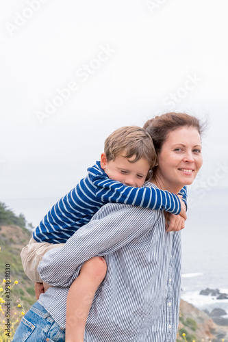 Attractive Young Mixed Race Mother and Son Hug Outdoors. Loving family having fun in nature park. Boy riding piggy ride on mothers back in summer day.
