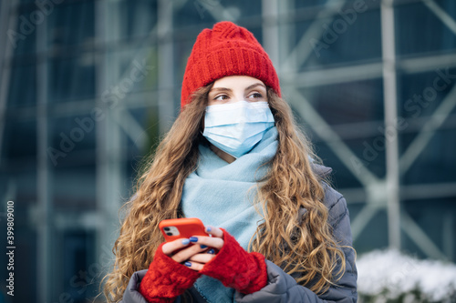 Joyful young woman texting on cellphone. Curly female standing on winter street. Caucasian beautiful curly female in mask browsing on smartphone while standing on street © uflypro