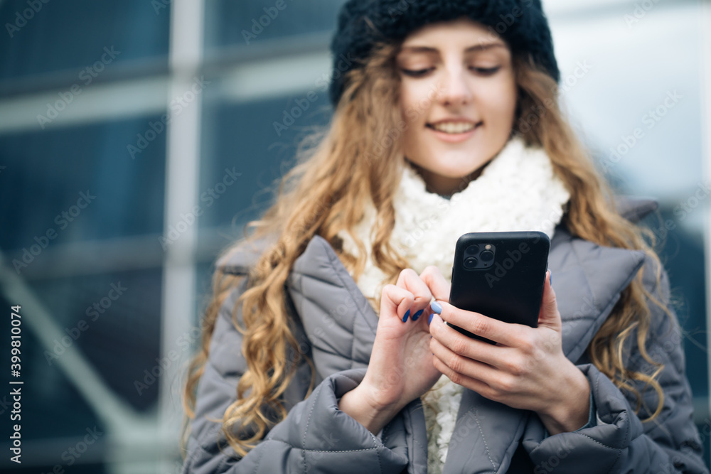 Joyful young female in good mood typing and scrolling on smartphone outdoors. Winter concept. Female using smartphone standing outside. Happy girl employee typing on a cellphone