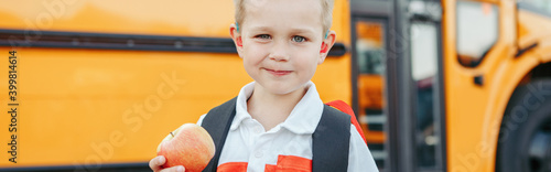 Caucasian boy student with backpack near yellow bus on first September day. Child kid eating apple at school yard outdoors. Education and back to school in Autumn Fall. Web banner header.
