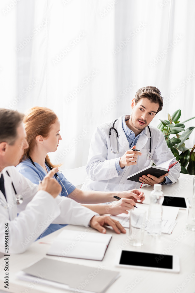 Doctor holding pen and notebook near colleagues, devices and papers on blurred foreground Doctor holding pen and notebook near colleagues, devices and papers on blurred foreground