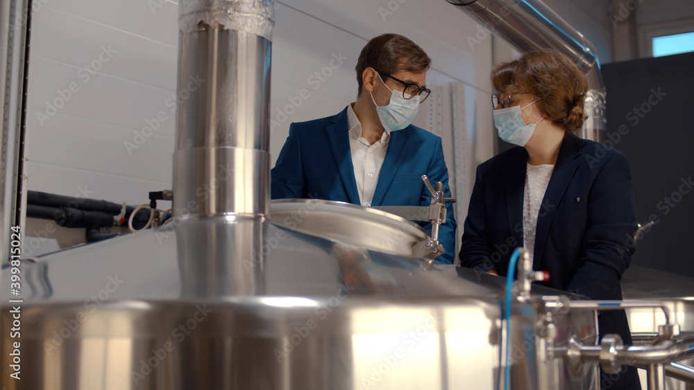Young male manager in suit and safety mask showing beer fermentation tank to female inspector