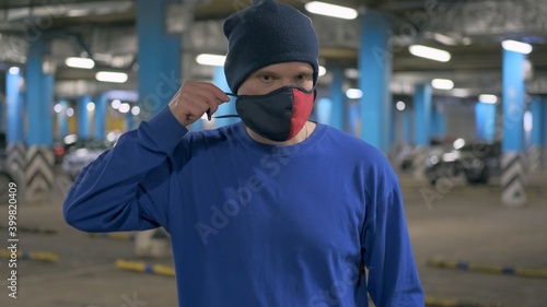Caucasian man in a hat stands in an underground Parking lot and puts on a red and black mask against the virus