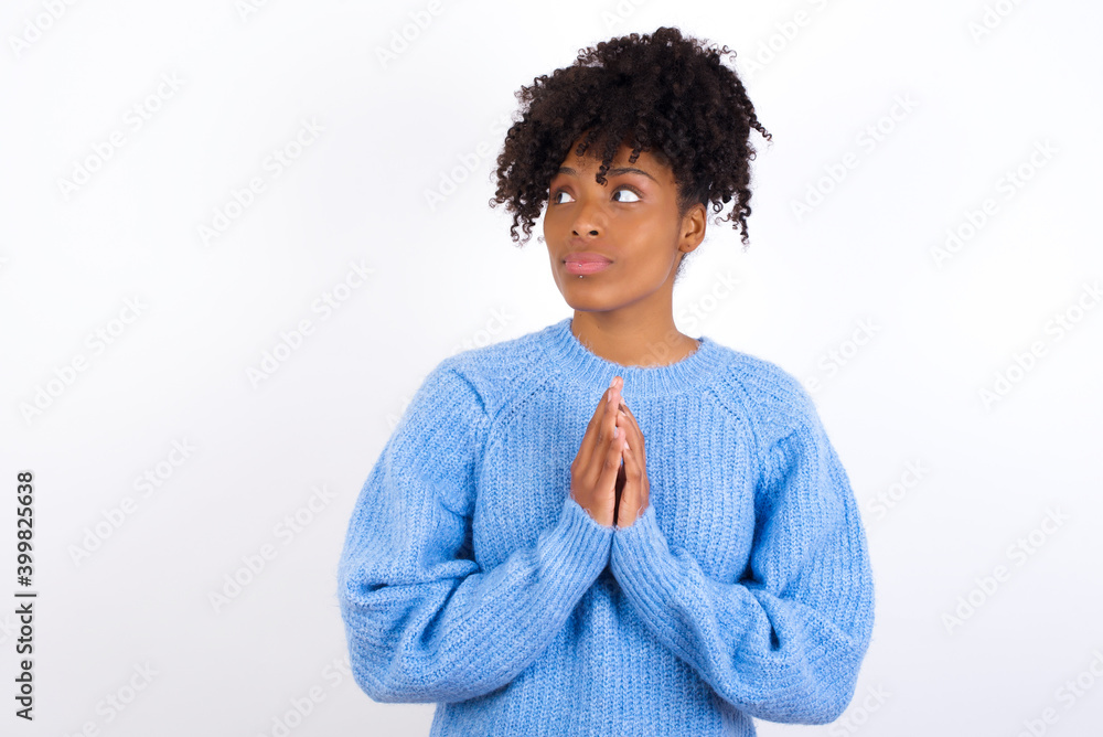 Charming cheerful oung beautiful African American woman wearing blue knitted sweater against white wall making up plan in mind holding hands together, setting up an idea. Look askance Charming cheerful oung beautiful African American woman wearing blue knitted sweater against white wall making up plan in mind holding hands together, setting up an idea. Look askance