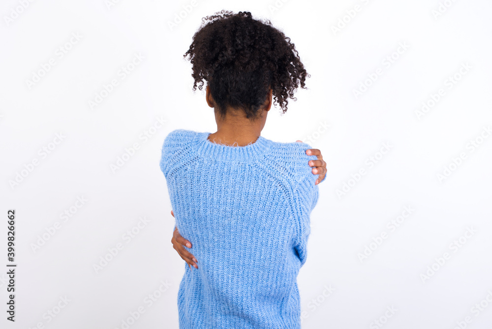 Young beautiful African American woman wearing blue knitted sweater against white wall hugging herself happy and positive from backwards. Self love and self care Young beautiful African American woman wearing blue knitted sweater against white wall hugging herself happy and positive from backwards. Self love and self care